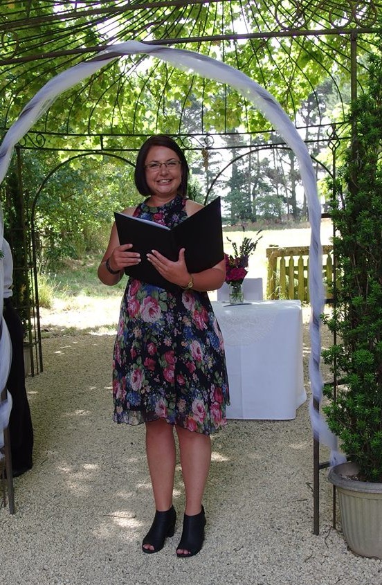 Danielle smiling under a garden arch at her first wedding, ceremony folder in hand
